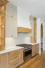 Kitchen with tasteful backsplash, gas stove, premium range hood, glass insert cabinets, and light wood-style floors