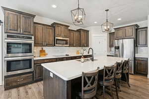 Kitchen featuring dark brown cabinets, tasteful backsplash, stainless steel appliances, a kitchen island with sink, and pendant lighting