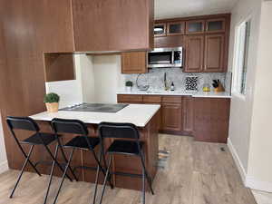 Kitchen featuring a kitchen breakfast bar, brown cabinets, backsplash, stainless steel microwave, and light wood-style floors