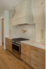Kitchen featuring custom exhaust hood, stainless steel gas range oven, light wood-style flooring, decorative backsplash, and light stone counters