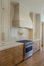 Kitchen featuring custom exhaust hood, high end stove, light wood-style floors, backsplash, and light stone counters