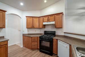 Kitchen with black range with gas cooktop, dishwasher, brown cabinetry, dark countertops, and light wood-style floors