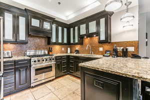 Kitchen with backsplash, luxury range, glass insert cabinets, hanging light fixtures, and light stone counters