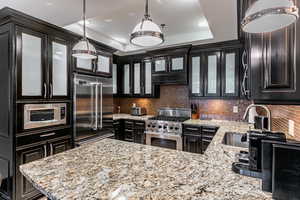 Kitchen featuring light stone countertops, glass insert cabinets, a tray ceiling, high end appliances, and tasteful backsplash
