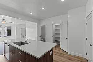 Kitchen with dark brown cabinetry, light wood-type flooring, light stone counters, recessed lighting, and an island with sink