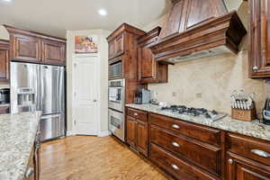 Kitchen featuring stainless steel appliances, backsplash, light wood-style flooring, light stone counters, and premium range hood