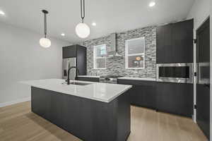 Kitchen featuring dark cabinets, wall chimney exhaust hood, pendant lighting, an island with sink, and light wood-type flooring