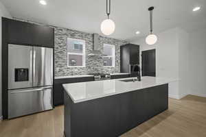 Kitchen featuring stainless steel appliances, light wood-style flooring, dark cabinets, wall chimney range hood, and pendant lighting