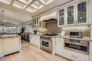 Kitchen featuring appliances with stainless steel finishes, premium range hood, glass insert cabinets, hanging light fixtures, and light stone countertops