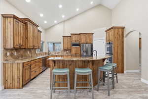 Kitchen featuring arched walkways, brown cabinets, decorative backsplash, light stone counters, and an island with sink