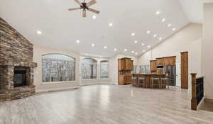 Living area with high vaulted ceiling, light wood-type flooring, a stone fireplace, a ceiling fan, and healthy amount of natural light