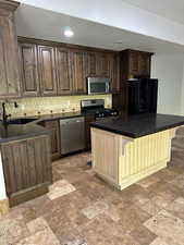 Second kitchen in the basement featuring dark brown cabinets, stainless steel appliances, a center island, backsplash, and a kitchen bar