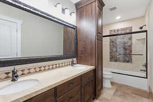 Guest bath featuring double vanity, combined bath / shower with glass door, stone tile flooring, and recessed lighting