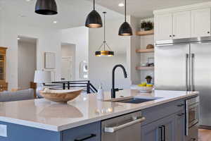Kitchen with light stone counters, open shelves, appliances with stainless steel finishes, pendant lighting, and a center island with sink