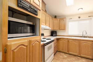 Kitchen featuring white electric stove, light countertops, stainless steel microwave, light brown cabinetry, and under cabinet range hood
