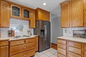 Kitchen featuring light countertops, backsplash, stainless steel fridge, light tile patterned flooring, and brown cabinetry