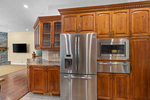 Kitchen with appliances with stainless steel finishes, brown cabinetry, dark stone counters, a warming drawer, and glass insert cabinets