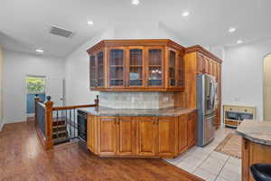 Kitchen featuring dark stone countertops, glass insert cabinets, stainless steel fridge with ice dispenser, brown cabinetry, and recessed lighting