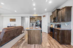 Kitchen with dark brown cabinets, decorative backsplash, black appliances, light stone countertops, and a kitchen island with sink