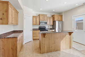 Kitchen with light tile patterned floors, dark stone counters, stainless steel appliances, a kitchen breakfast bar, and a kitchen island with sink