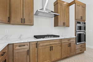 Kitchen with wall chimney exhaust hood, appliances with stainless steel finishes, brown cabinetry, and light stone countertops