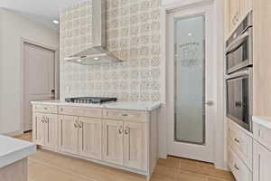 Kitchen with light brown cabinetry, wall chimney exhaust hood, stainless steel appliances, light stone countertops, and light tile patterned floors