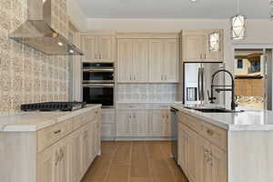 Kitchen featuring light brown cabinetry, backsplash, wall chimney range hood, hanging light fixtures, and a kitchen island with sink