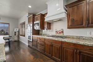 Kitchen with custom exhaust hood, dark wood-type flooring, appliances with stainless steel finishes, light stone counters, and a textured ceiling