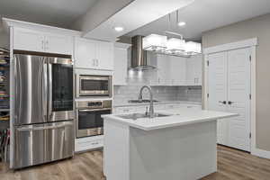 Kitchen featuring stainless steel appliances, wall chimney exhaust hood, backsplash, light wood finished floors, and white cabinets