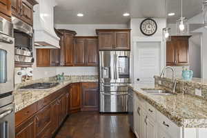Kitchen featuring appliances with stainless steel finishes, light stone counters, premium range hood, decorative light fixtures, and a textured ceiling