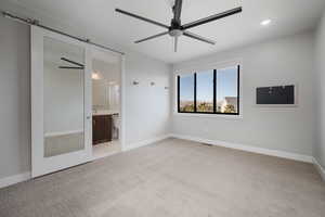 Unfurnished bedroom featuring light colored carpet, ceiling fan, a barn door, ensuite bath, and recessed lighting