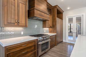 Kitchen with high end stainless steel range, brown cabinetry, custom range hood, french doors, and dark wood finished floors