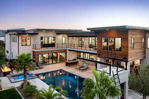 Back of house at dusk featuring a pool with connected hot tub, a patio area, a balcony, and stairway
