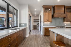 Kitchen featuring brown cabinets, light stone countertops, light wood-style flooring, tasteful backsplash, and glass insert cabinets