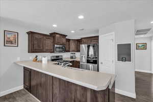 Kitchen with dark brown cabinetry, stainless steel appliances, light countertops, a peninsula, and dark wood-style floors