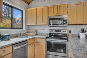 Kitchen featuring stainless steel appliances, light stone counters, light brown cabinets, vaulted ceiling, and wood walls