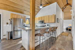 Kitchen featuring wooden walls, stainless steel appliances, dark wood-type flooring, a kitchen breakfast bar, and light stone countertops