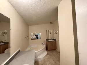 Primary bathroom with a textured wall, two vanities, light wood-type flooring, a garden tub, and a textured ceiling