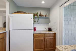 Kitchen featuring freestanding refrigerator, open shelves, brown cabinetry, light stone countertops, and decorative backsplash
