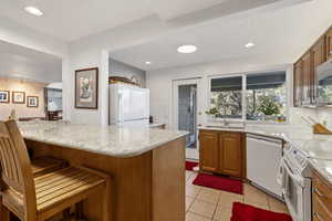 Kitchen featuring tasteful backsplash, brown cabinetry, light tile patterned floors, a kitchen breakfast bar, and white appliances