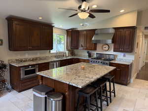 Kitchen with dark brown cabinets, stone tile floors, wall chimney exhaust hood, recessed lighting, and vaulted ceiling