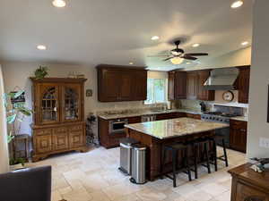 Kitchen with dark brown cabinets, wall chimney exhaust hood, a breakfast bar, appliances with stainless steel finishes, and light stone counters