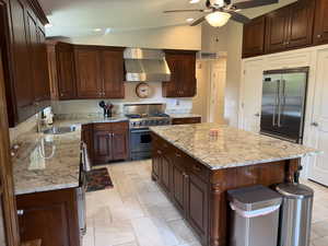 Kitchen featuring wall chimney range hood, high end appliances, light stone counters, vaulted ceiling, and a kitchen island