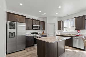 Kitchen featuring stainless steel appliances, dark brown cabinetry, light stone countertops, light wood-type flooring, and recessed lighting