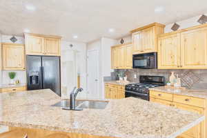 Kitchen featuring light brown cabinetry, black appliances, decorative backsplash, recessed lighting, and an island with sink