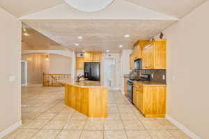 Kitchen with arched walkways, black appliances, light stone countertops, decorative backsplash, and a kitchen island with sink