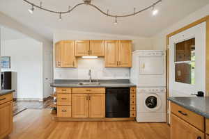 Kitchen with rail lighting, dark countertops, light wood finished floors, black dishwasher, and stacked washing machine and dryer