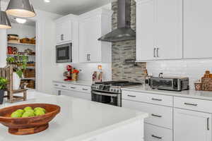 Kitchen with wall chimney range hood, white cabinetry, tasteful backsplash, stainless steel gas range, and light stone counters