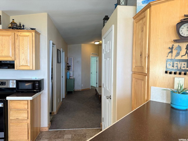 Kitchen with stainless steel electric range oven, light brown cabinetry, black microwave, exhaust hood, and dark colored carpet