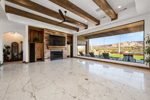 Unfurnished living room featuring beamed ceiling, light marble finish flooring, ceiling fan, arched walkways, and a fireplace